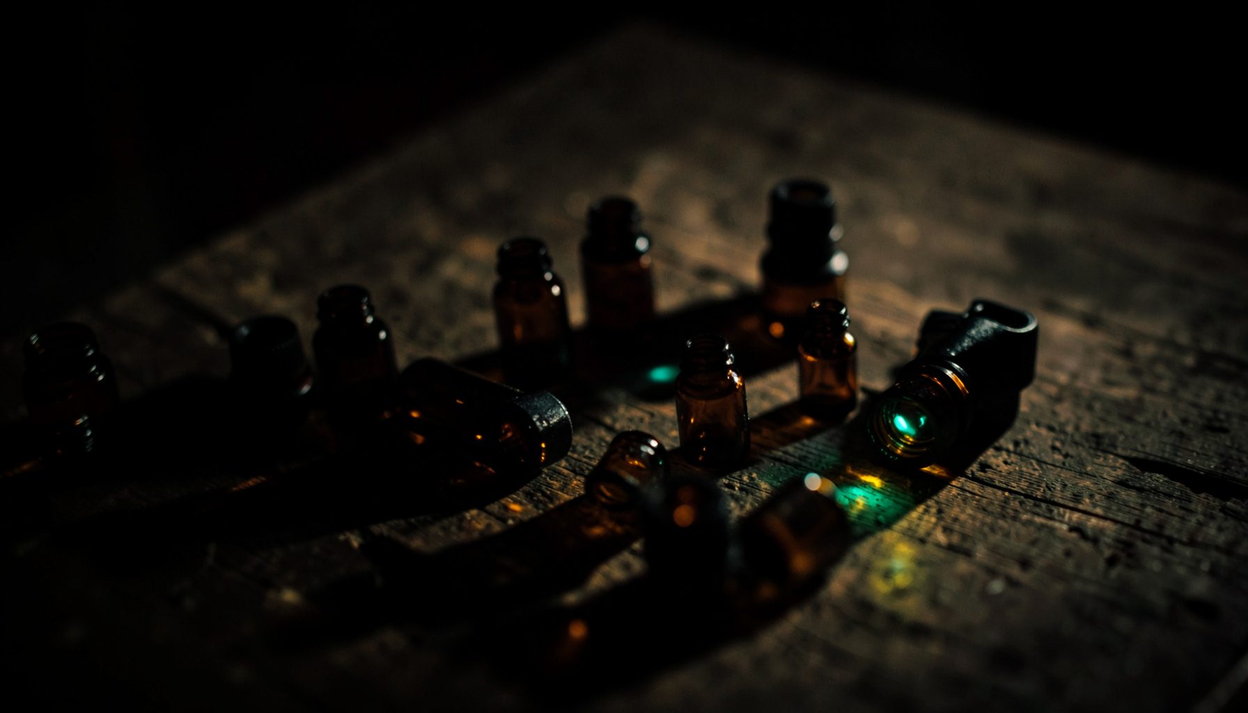 Dark, low-key photograph of small amber glass essential oil bottles scattered on a weathered wooden surface, deep shadows and warm golden highlights